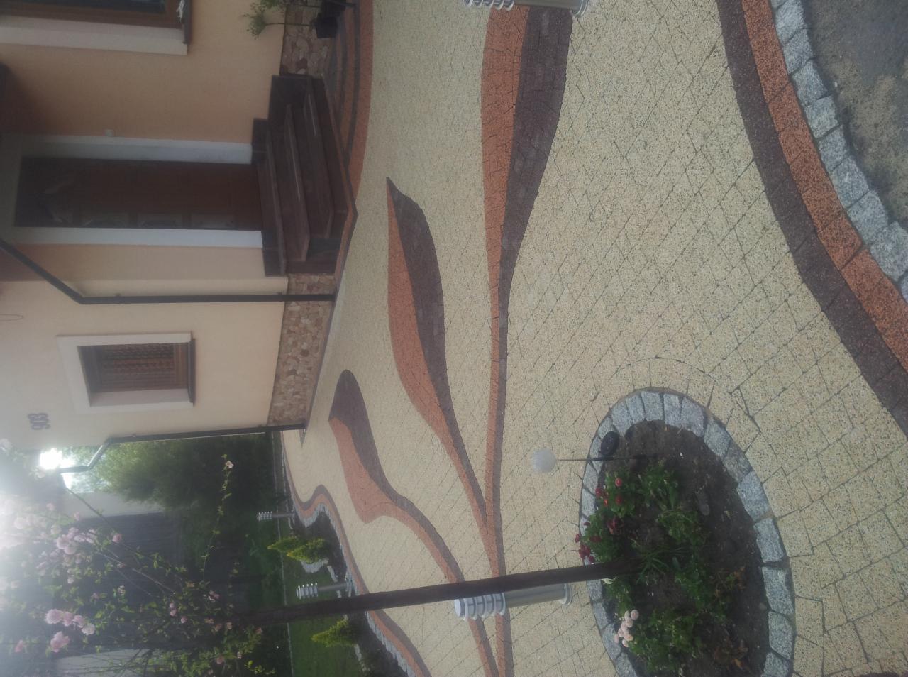 Ułożona kostka brukowa w odcieniach beżu, brązu i szarości tworząca wzór liści na podjeździe przed domem z elementami małej architektury ogrodowej.