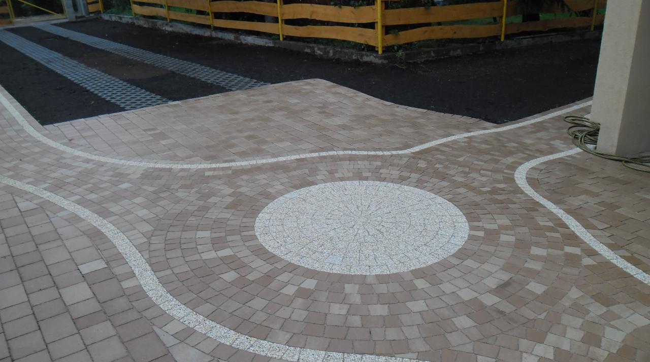 Ułożona kostka brukowa w odcieniach beżu z białymi akcentami tworzącymi wzór koła i łuków, widoczna część podjazdu z ażurowymi płytami, żółte drewniane ogrodzenie w tle.