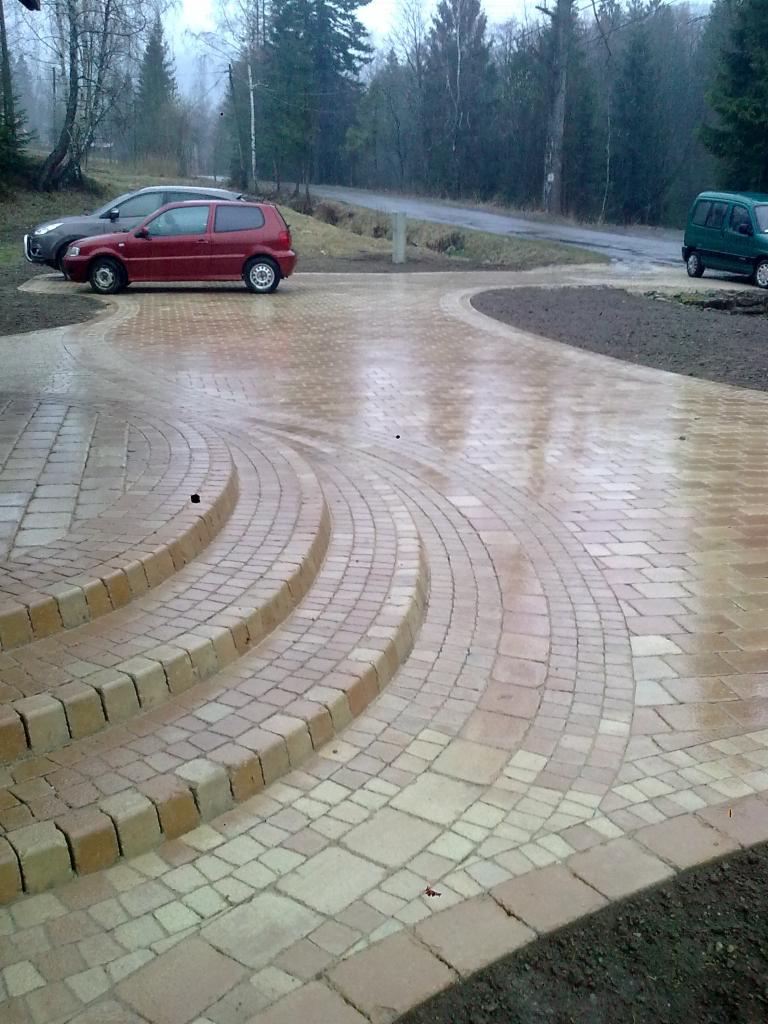 Ułożona kostka brukowa w kształcie łuków tworząca podjazd i schody, widoczne dwa samochody na tle drzew