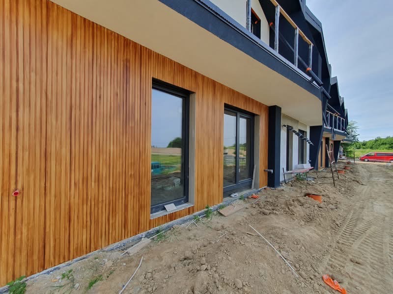 Nowoczesny budynek z elewacją z drewnianych paneli, czarnymi ramami okiennymi i balkonami. Widoczne prace ziemne wokół budynku.