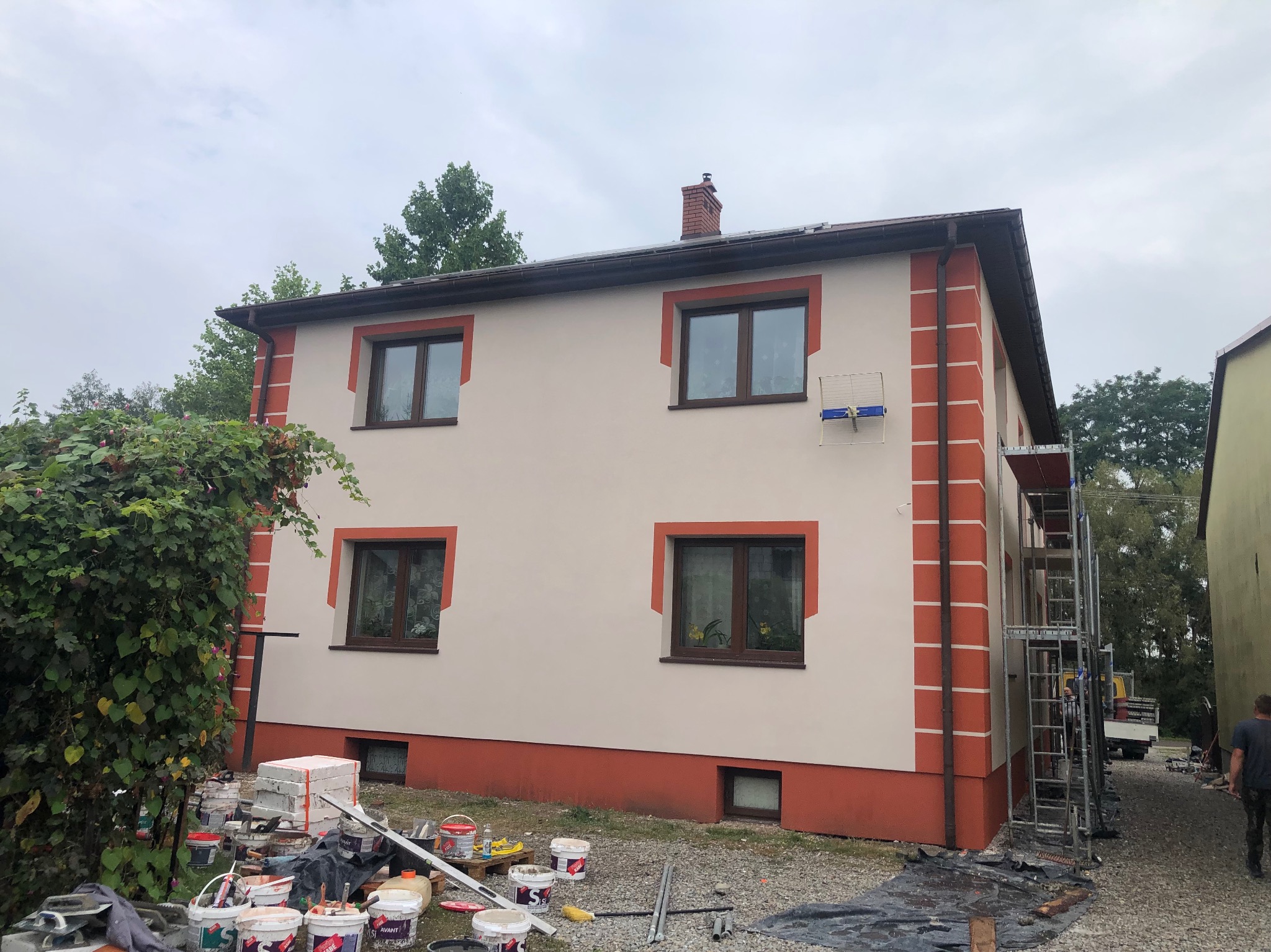 Dwukondygnacyjny dom z elewacją w odcieniach beżu i czerwieni, z widocznymi rusztowaniami i materiałami budowlanymi. Świeżo pomalowana fasada.