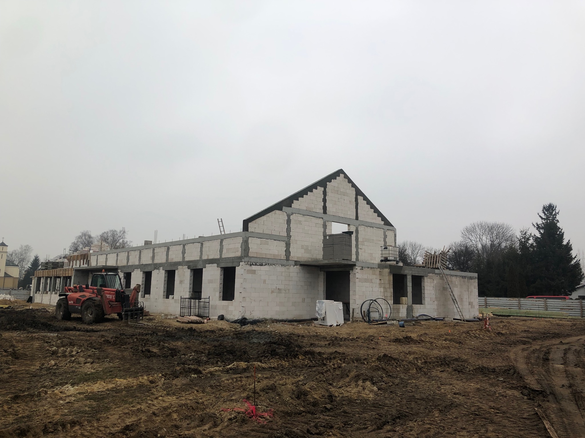 Budowa domu z białych bloczków, widoczny częściowo postawiony budynek z otworami okiennymi i sprzęt budowlany na placu budowy w pochmurny dzień.