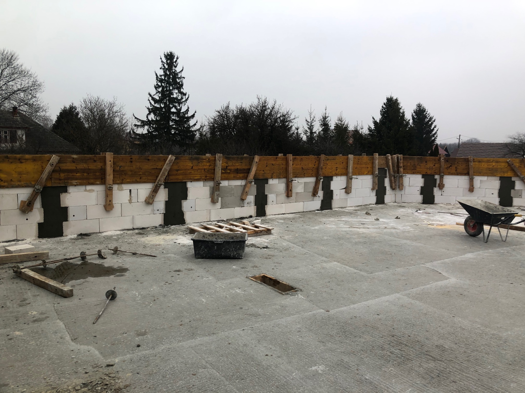 Wznoszenie ścian z bloczków betonowych z drewnianym szalunkiem. Widoczny wózek budowlany, narzędzia i elementy konstrukcyjne na betonowej płycie fundamentowej.