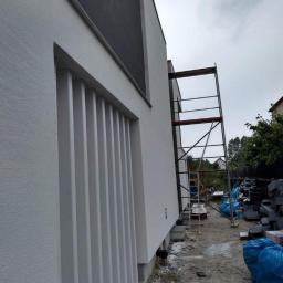 Fhu Izabela - Nowoczesny budynek z jasną elewacją o fakturze baranka, z elementami dekoracyjnymi w postaci pionowych listew przy oknie, widoczne rusztowanie i materiały budowlane w tle.