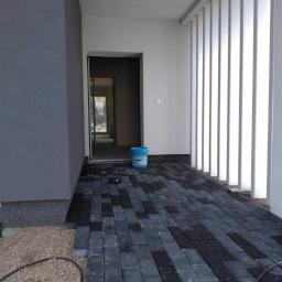 Fhu Izabela - Układanie szarej kostki brukowej przed wejściem do budynku z szarą elewacją, widoczny fragment podmurówki i wiadro z farbą w tle.