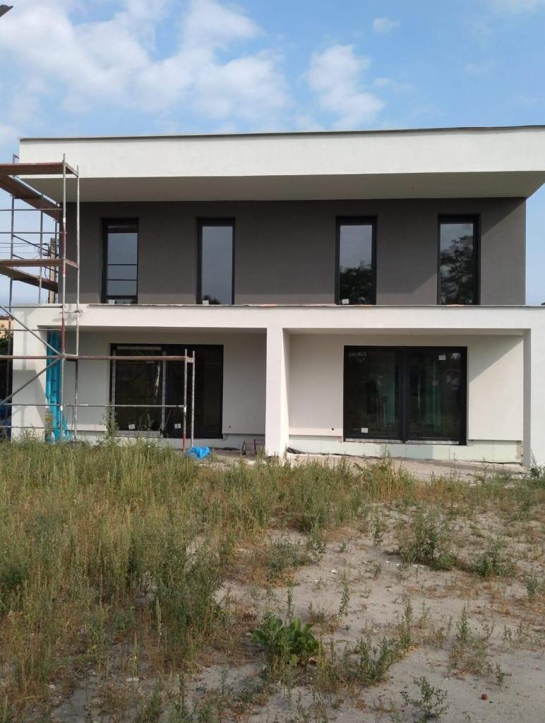 Dwukondygnacyjny dom w trakcie wykańczania elewacji, widoczne rusztowania i świeżo nałożony tynk w odcieniach szarości i bieli, zarośnięty teren przed budynkiem.