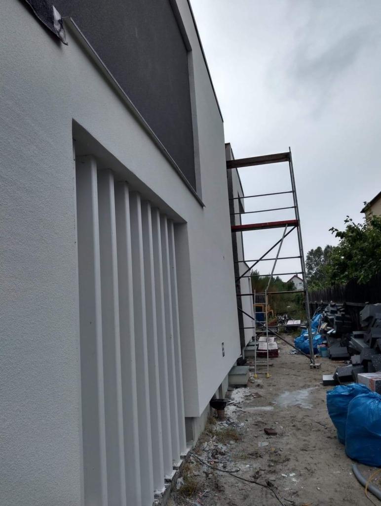 Nowoczesny budynek z jasną elewacją o fakturze baranka, z elementami dekoracyjnymi w postaci pionowych listew przy oknie, widoczne rusztowanie i materiały budowlane w tle.