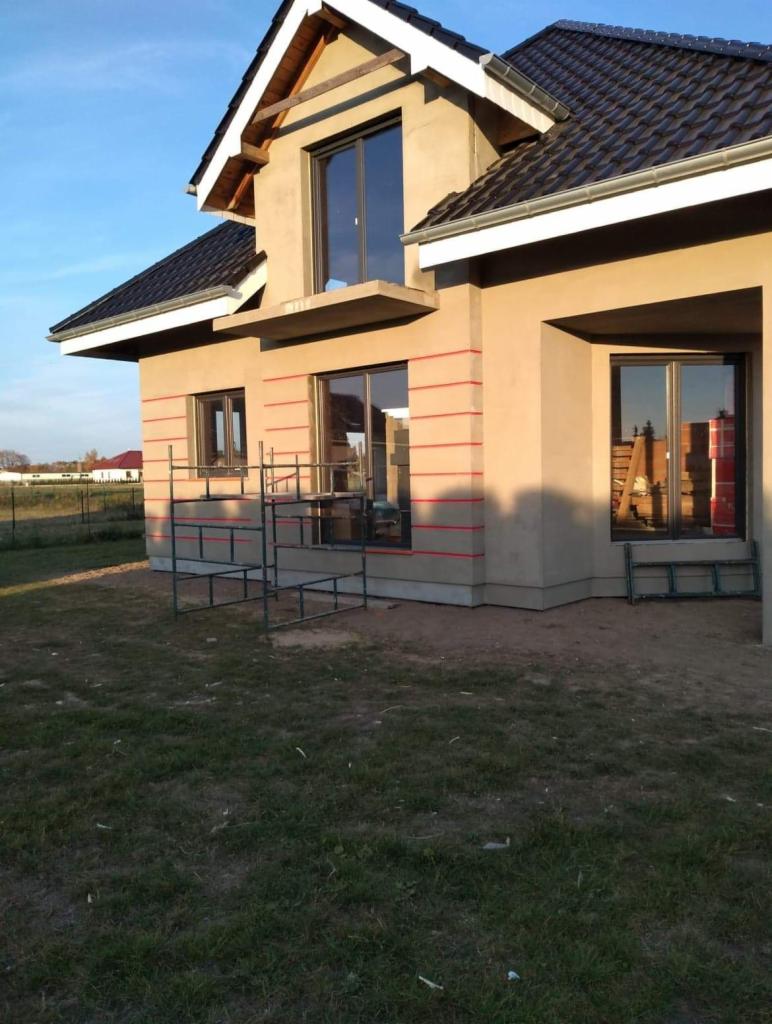 Dom jednorodzinny w trakcie nakładania warstwy elewacyjnej, widoczne rusztowanie i czerwone linie wyznaczające poziomy, dach z ciemnej dachówki.