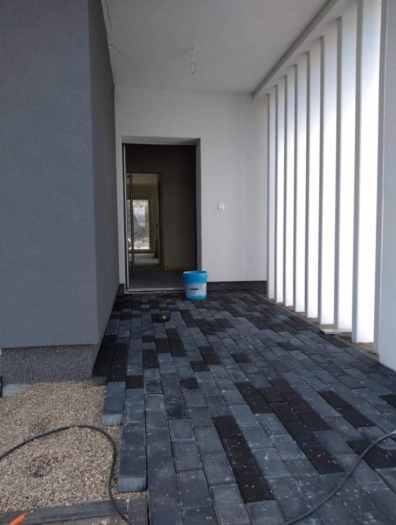 Układanie szarej kostki brukowej przed wejściem do budynku z szarą elewacją, widoczny fragment podmurówki i wiadro z farbą w tle.