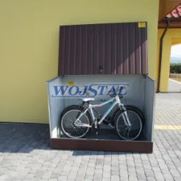 Metalowy schowek na rowery z otwartymi drzwiami, z rowerem w środku, na tle żółtej ściany budynku i kostki brukowej.