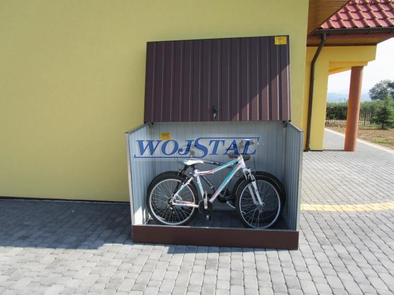 Metalowy schowek na rowery z otwartymi drzwiami, z rowerem w środku, na tle żółtej ściany budynku i kostki brukowej.