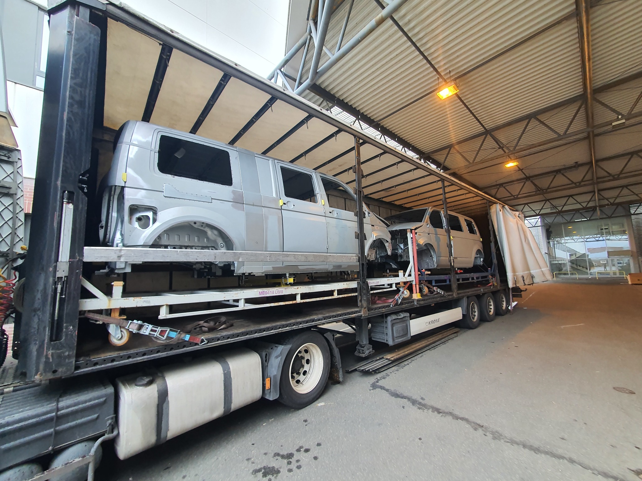 Ciężarówka z otwartą naczepą przewożąca dwa szare, niekompletne nadwozia busów, zabezpieczone pasami transportowymi.