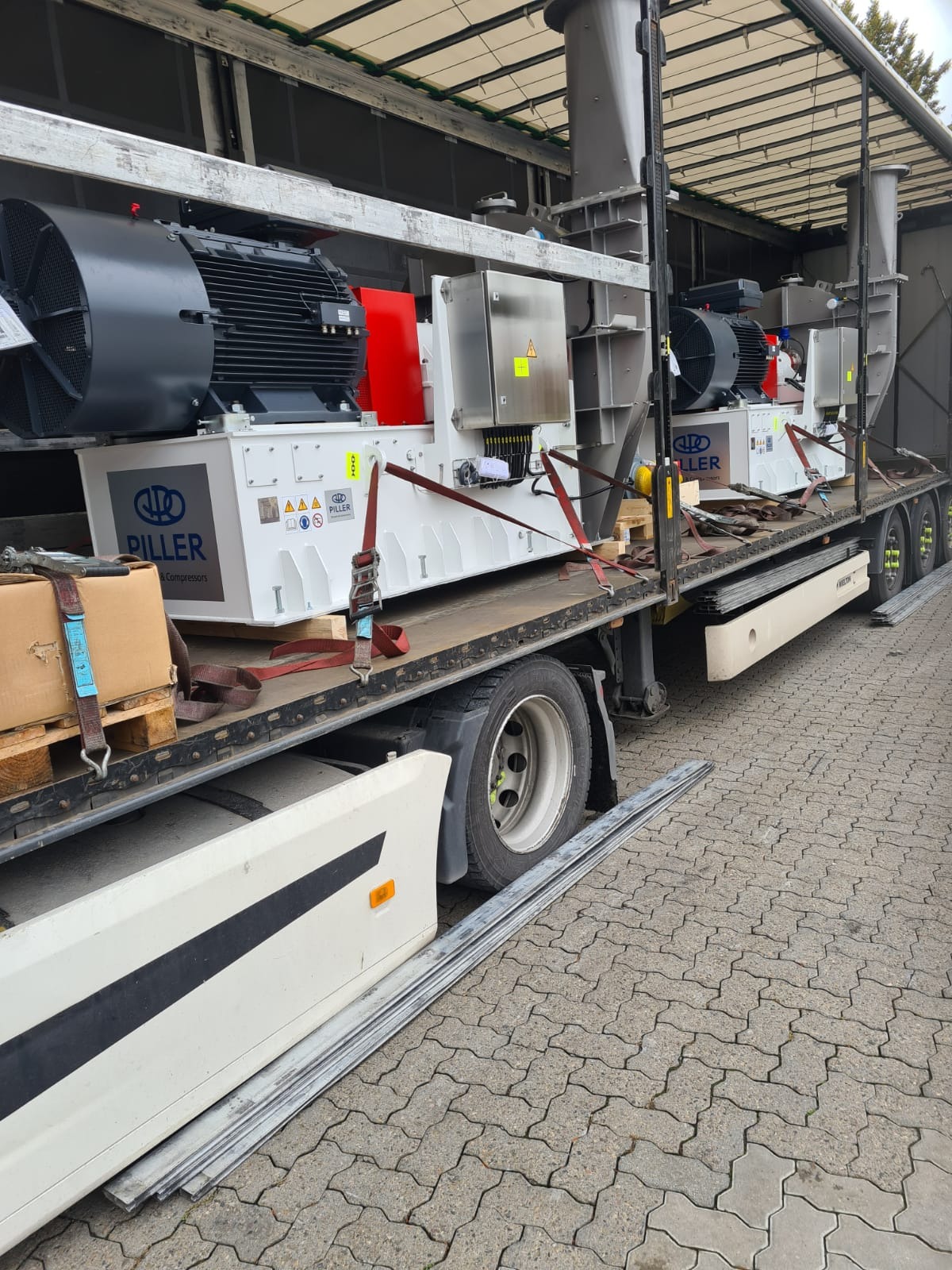 Ciężarówka z naczepą przewozi dwa duże, białe urządzenia przemysłowe marki Piller, zabezpieczone czerwonymi pasami transportowymi.