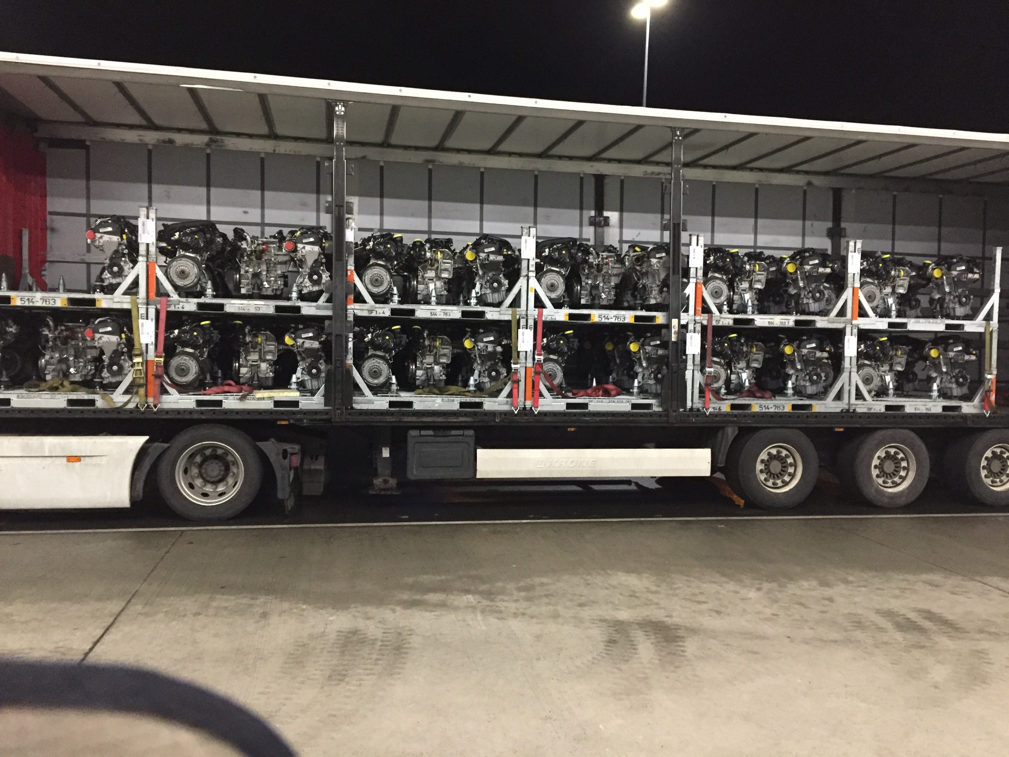 Ciężarówka z naczepą załadowana silnikami samochodowymi na metalowych regałach, zabezpieczonych pasami transportowymi; widok z boku, oświetlenie nocne.