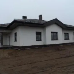 Niedawno wybudowany, parterowy dom jednorodzinny z ciemną dachówką, jasną elewacją i dwoma kominami, otoczony ziemią przygotowaną pod zagospodarowanie terenu.