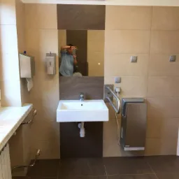 Łazienka publiczna z umywalką, suszarką do rąk i dużymi, beżowo-brązowymi płytkami, z odbiciem fotografa w lustrze.