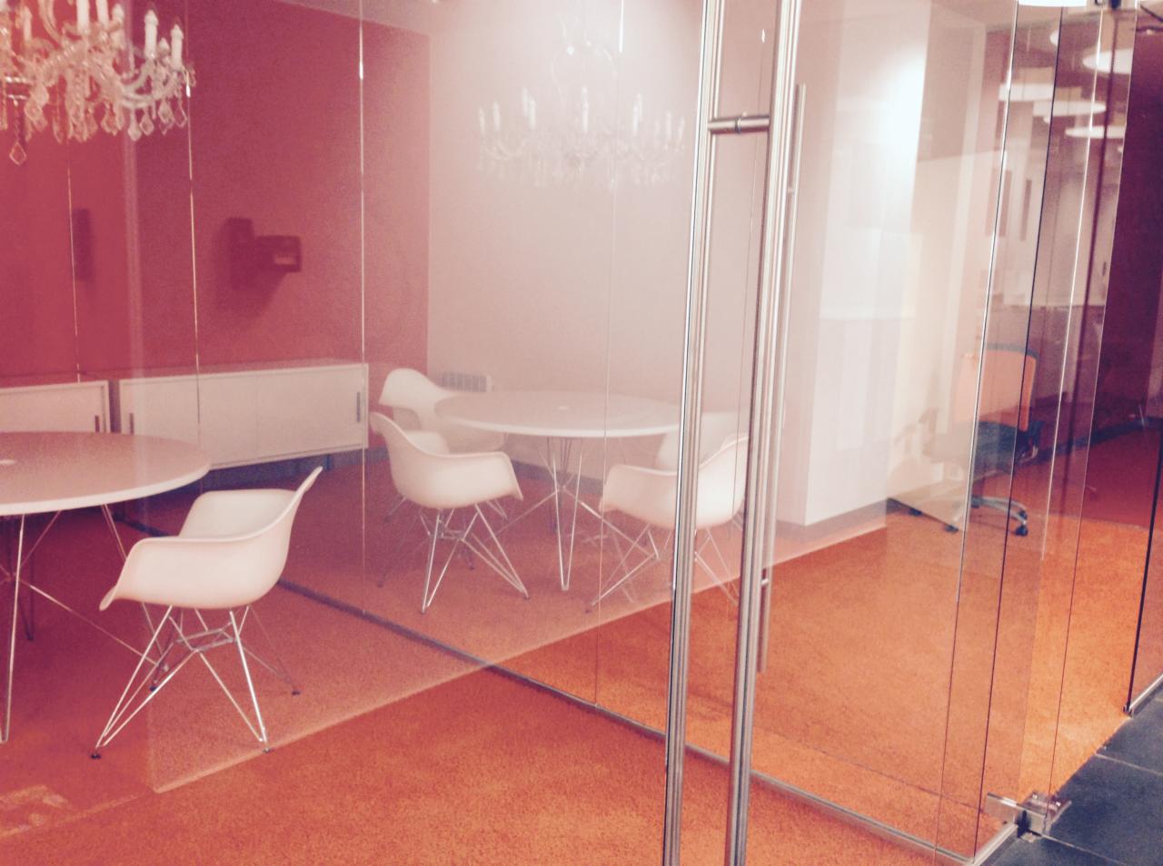 Szklana ściana działowa z metalowymi okuciami oddzielająca nowoczesne biuro z okrągłymi stołami i białymi krzesłami Eames, widoczne żyrandole i pomarańczowa wykładzina podłogowa.