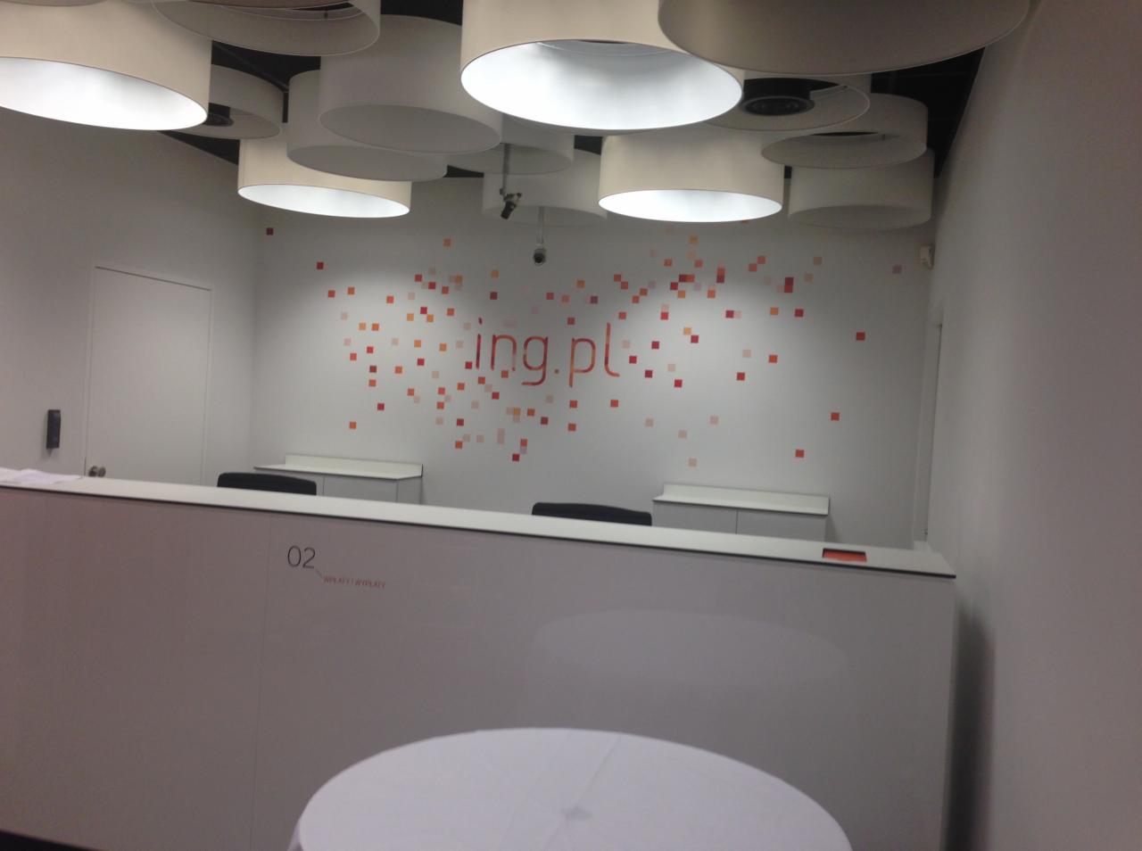 Recepcja z białym blatem, dekoracyjną ścianą z logo i okrągłymi lampami sufitowymi.
