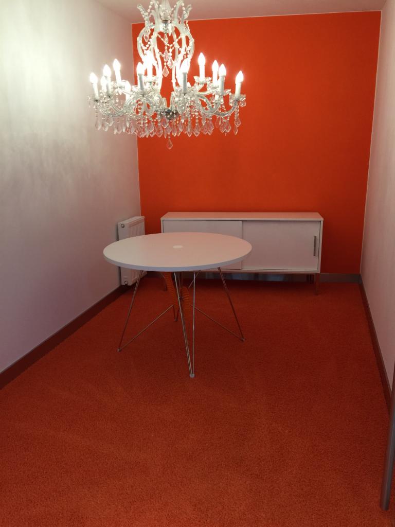 Wnętrze pokoju z pomarańczową ścianą, białym kredensem, okrągłym stołem na metalowych nogach i kryształowym żyrandolem.