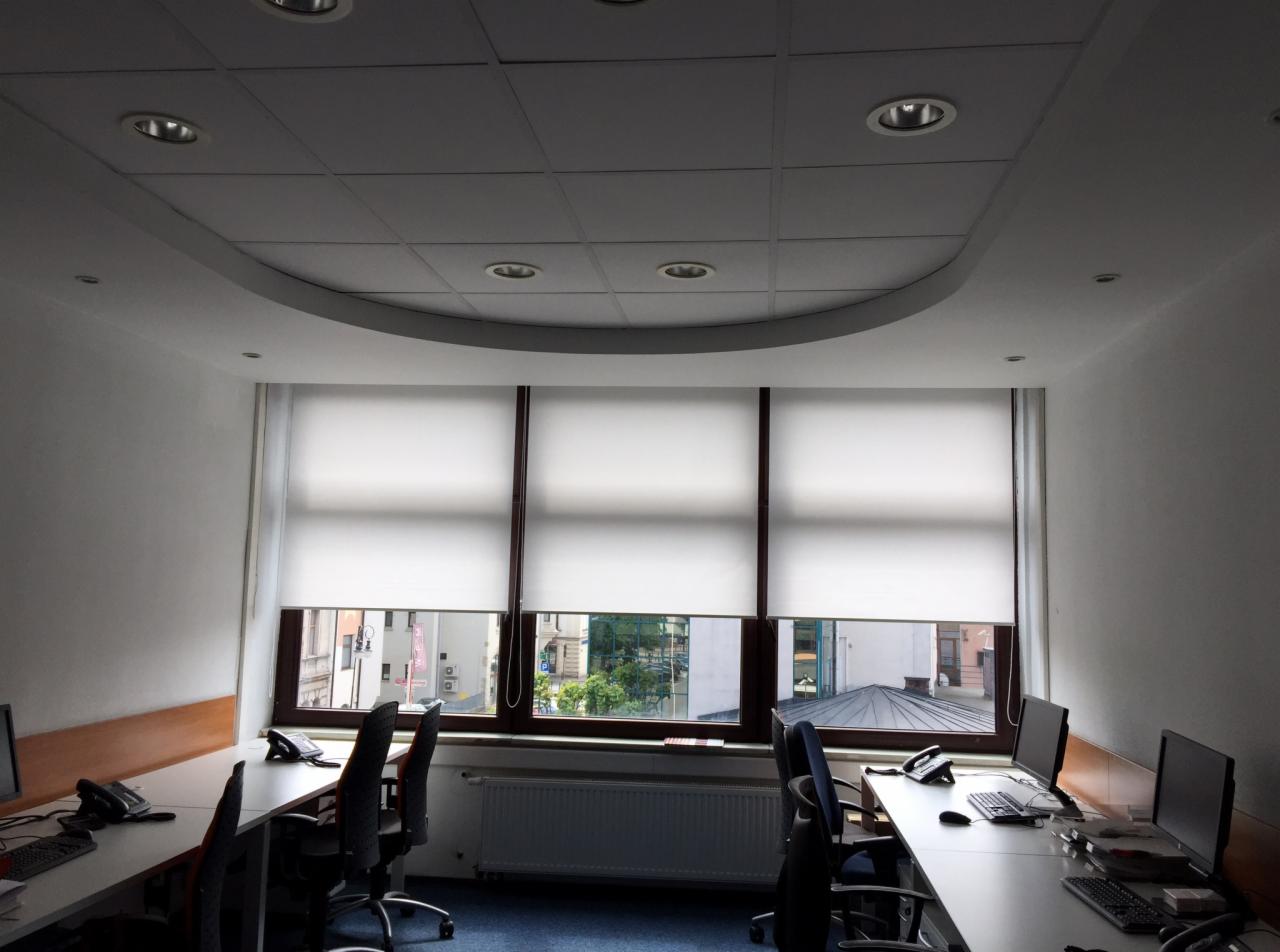 Biuro z trzema oknami z białymi roletami, rzędem biurek z komputerami i telefonami oraz sufit podwieszany z okrągłymi lampami.