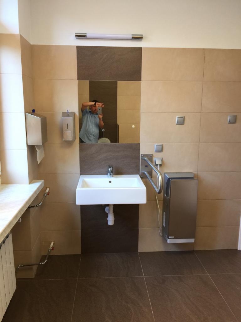 Łazienka publiczna z umywalką, suszarką do rąk i dużymi, beżowo-brązowymi płytkami, z odbiciem fotografa w lustrze.