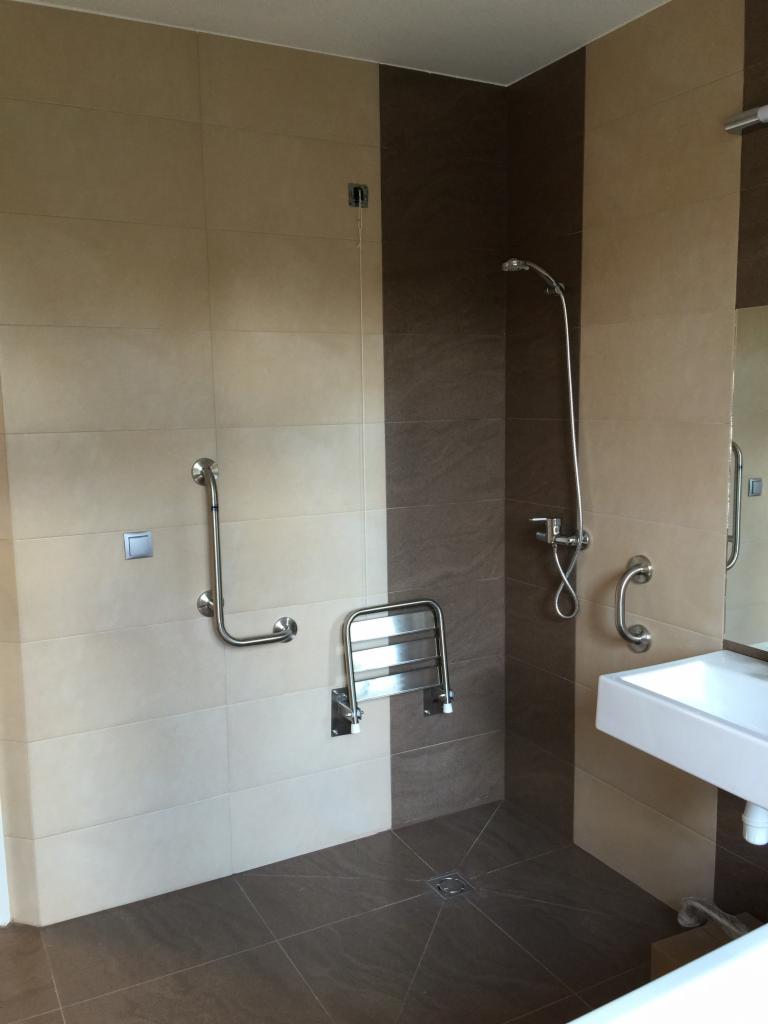Wykończona łazienka z brązowo-beżowymi płytkami, prysznicem bez brodzika z siedziskiem i uchwytami dla osób z niepełnosprawnościami, umywalką oraz odpływem liniowym w podłodze.