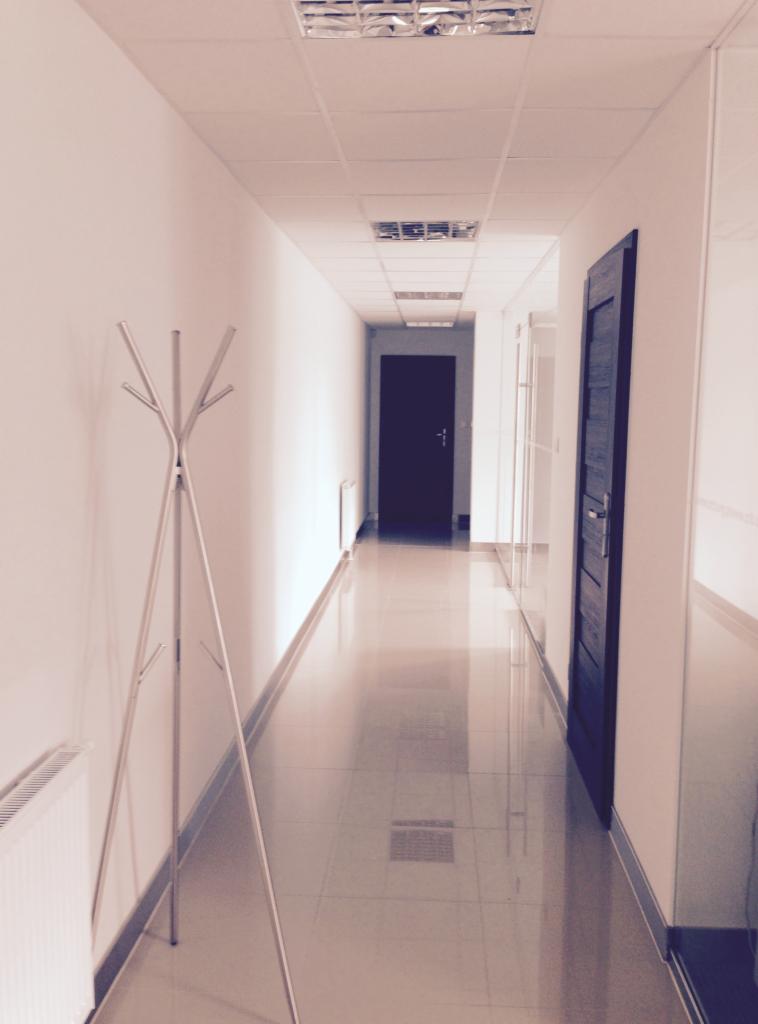 Długi korytarz biurowy z błyszczącą podłogą z płytek, widoczne drzwi, grzejnik i wieszak na ubrania.