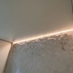 Ściana z dekoracyjną fakturą oświetlona listwą LED pod sufitem, widoczne płyty kartonowo-gipsowe przy ścianie.