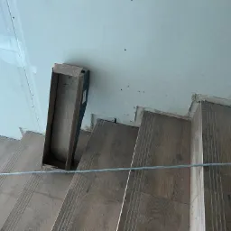 Montaż płyty karton-gips na ścianie przy schodach z drewnopodobnymi stopniami, widoczna listwa przypodłogowa i skrzynka narzędziowa.