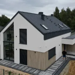 Nowoczesny dom z grafitowym dachem, oknami z żaluzjami zewnętrznymi i elementami drewnianej elewacji. Widok z góry na posesję z kostką brukową.