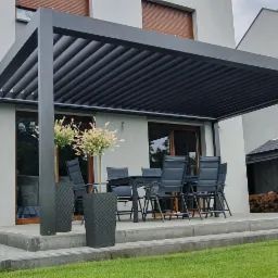 Nowoczesna pergola Selt SB400 z regulowanymi lamelami, zacieniająca taras z meblami ogrodowymi przy domu z jasną elewacją.