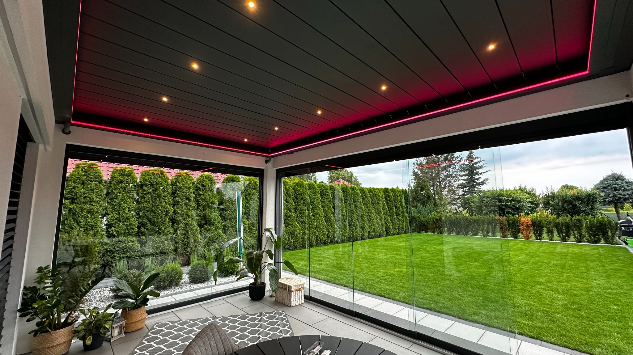 Nowoczesna pergola metalowa z oświetleniem LED, widok z wnętrza na zadbany ogród. Szklane ściany przesuwne zapewniają panoramiczny widok.
