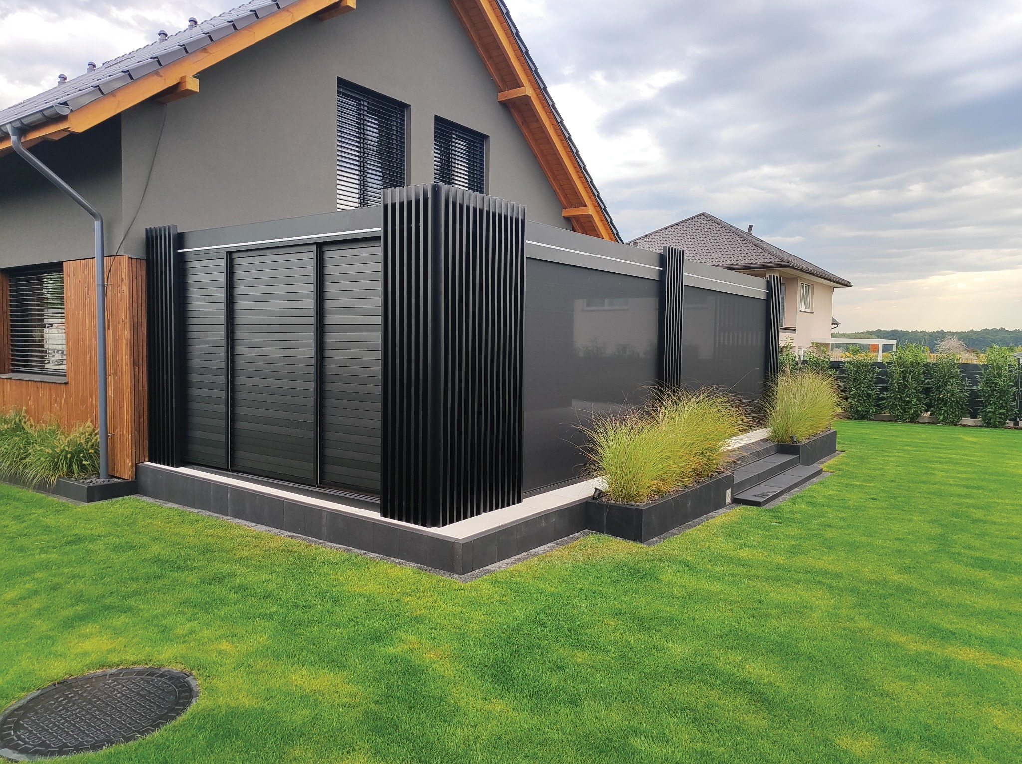 Nowoczesna, czarna pergola metalowa z przesuwnymi panelami, przylegająca do domu z szarą elewacją. Elementy dekoracyjne z trawy ozdobnej. Widok na zadbany trawnik.