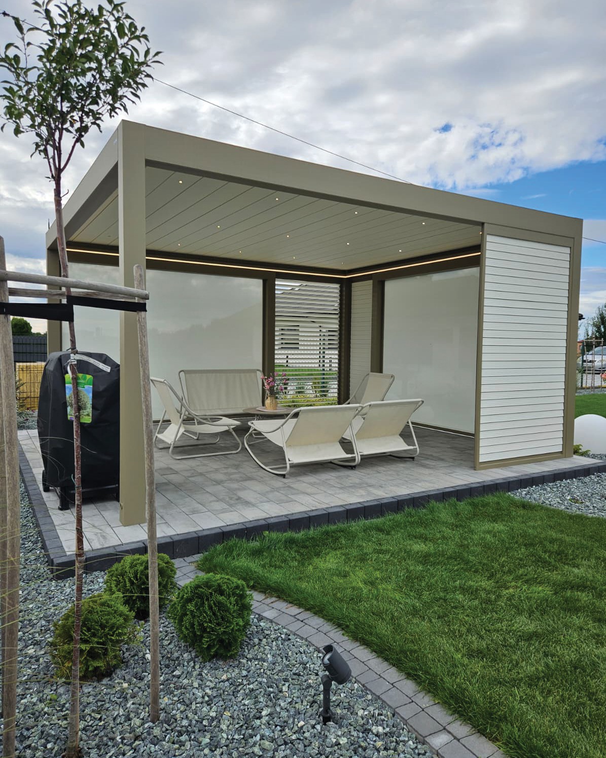 Nowoczesna pergola metalowa z oświetleniem LED i meblami ogrodowymi na tarasie z kostki brukowej, otoczona zielenią i ozdobnym żwirem.