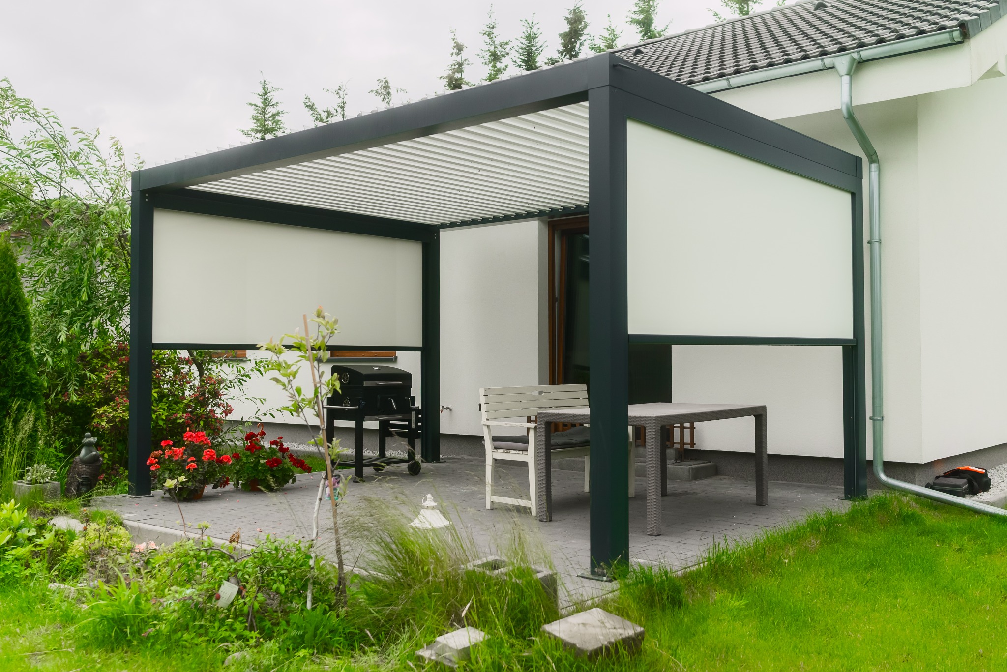 Nowoczesna pergola metalowa z roletami przeciwsłonecznymi, stół z krzesłami i grill na tarasie z kostki brukowej, otoczona zielenią i kwiatami.
