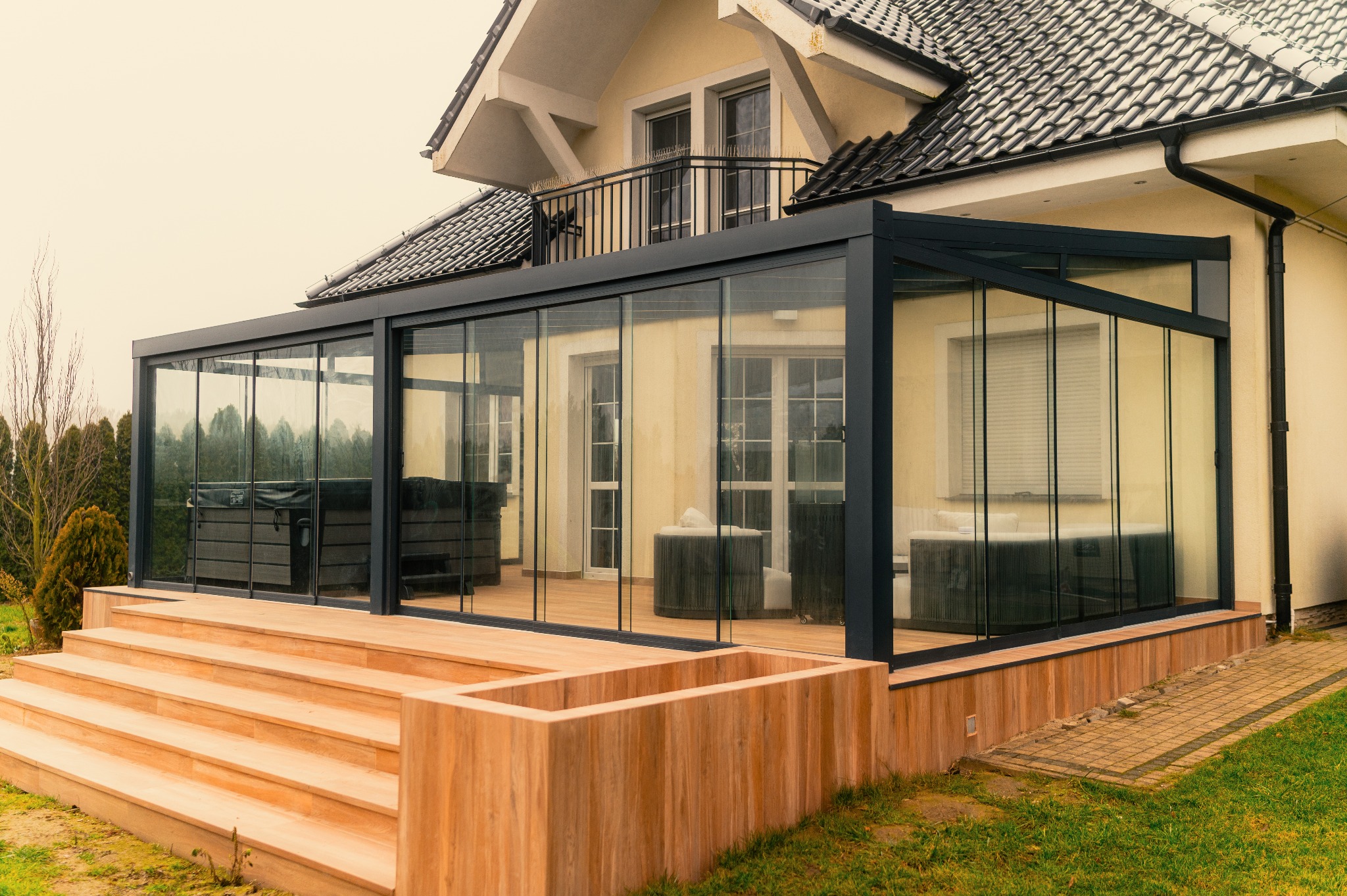 Nowoczesna, przeszklona pergola z czarną ramą, przylegająca do domu z balkonem. Widoczne drewniane schody i jacuzzi wewnątrz. Jasne, naturalne światło.