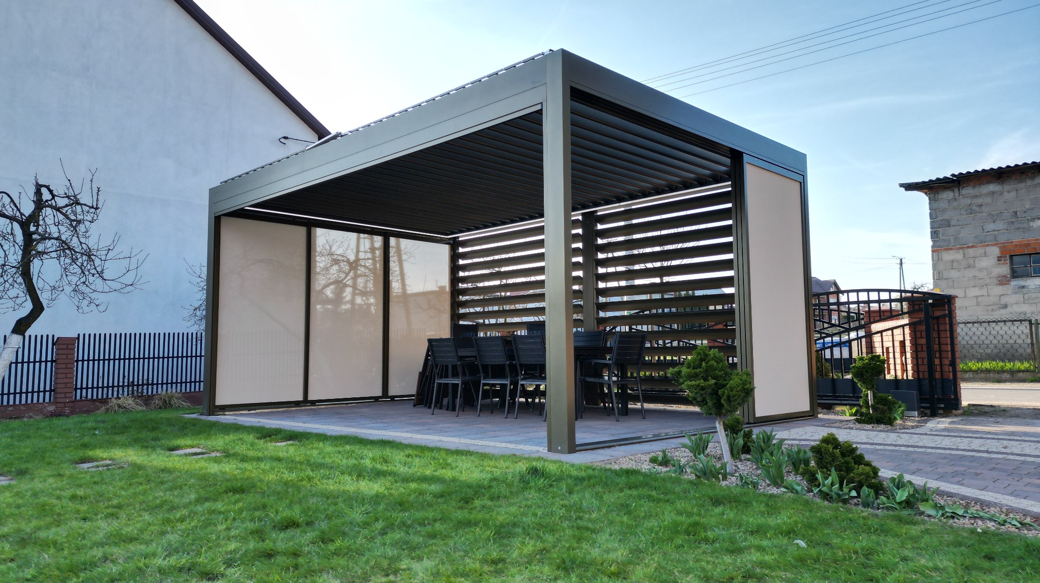 Nowoczesna, metalowa pergola z żaluzjowym dachem i rolowanymi bokami, ustawiona na kostce brukowej w ogrodzie. Wewnątrz zestaw mebli ogrodowych.