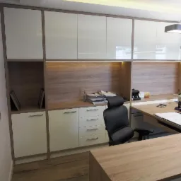 Nowoczesne biuro z zabudową meblową: białe, lakierowane szafki górne i dolne z drewnianym dekorem, oświetlenie LED pod szafkami, biurko z blatem imitującym jasne drewno, czarne krzesło obrotowe.