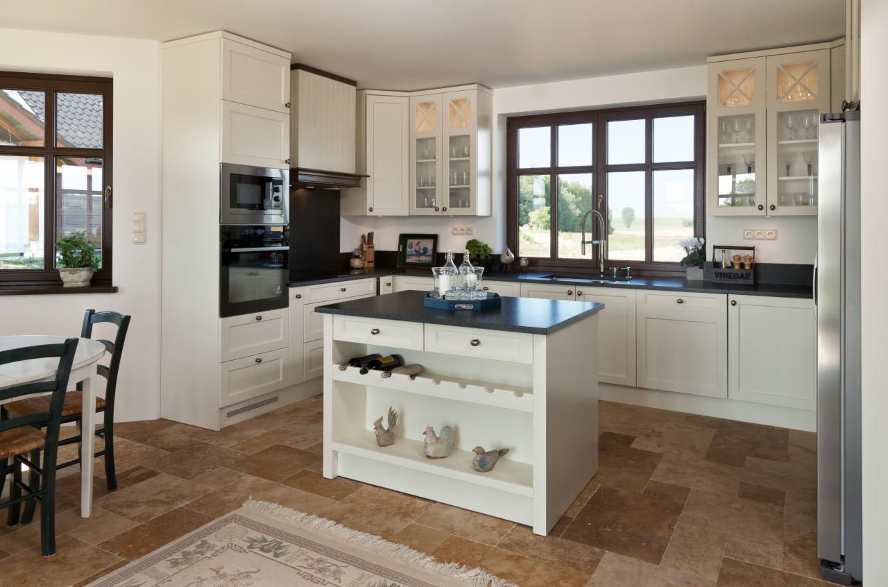 Jasna kuchnia z wyspą, czarnym blatem i brązowymi ramami okiennymi, płytki podłogowe w odcieniach brązu, widok na zieleń za oknem.