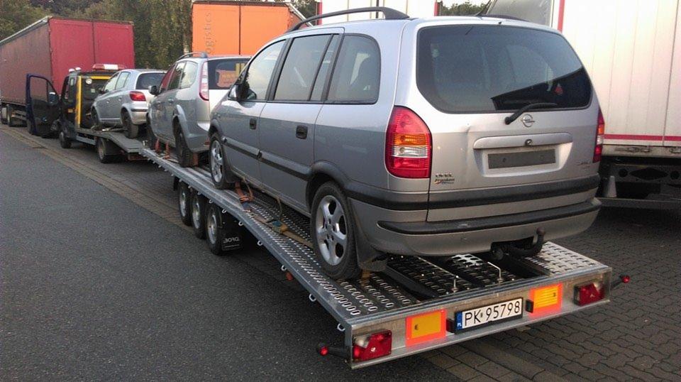 Srebrny Opel Zafira zabezpieczony pasami na platformie lawety, z widoczną tablicą rejestracyjną, w tle inne samochody i ciężarówki.