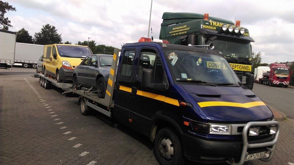 Niebieska laweta z żółtymi pasami transportuje szary samochód osobowy i żółty van na parkingu, w tle widoczne inne ciężarówki, w tym zielona ciężarówka z napisem 'KOSZALIN'.