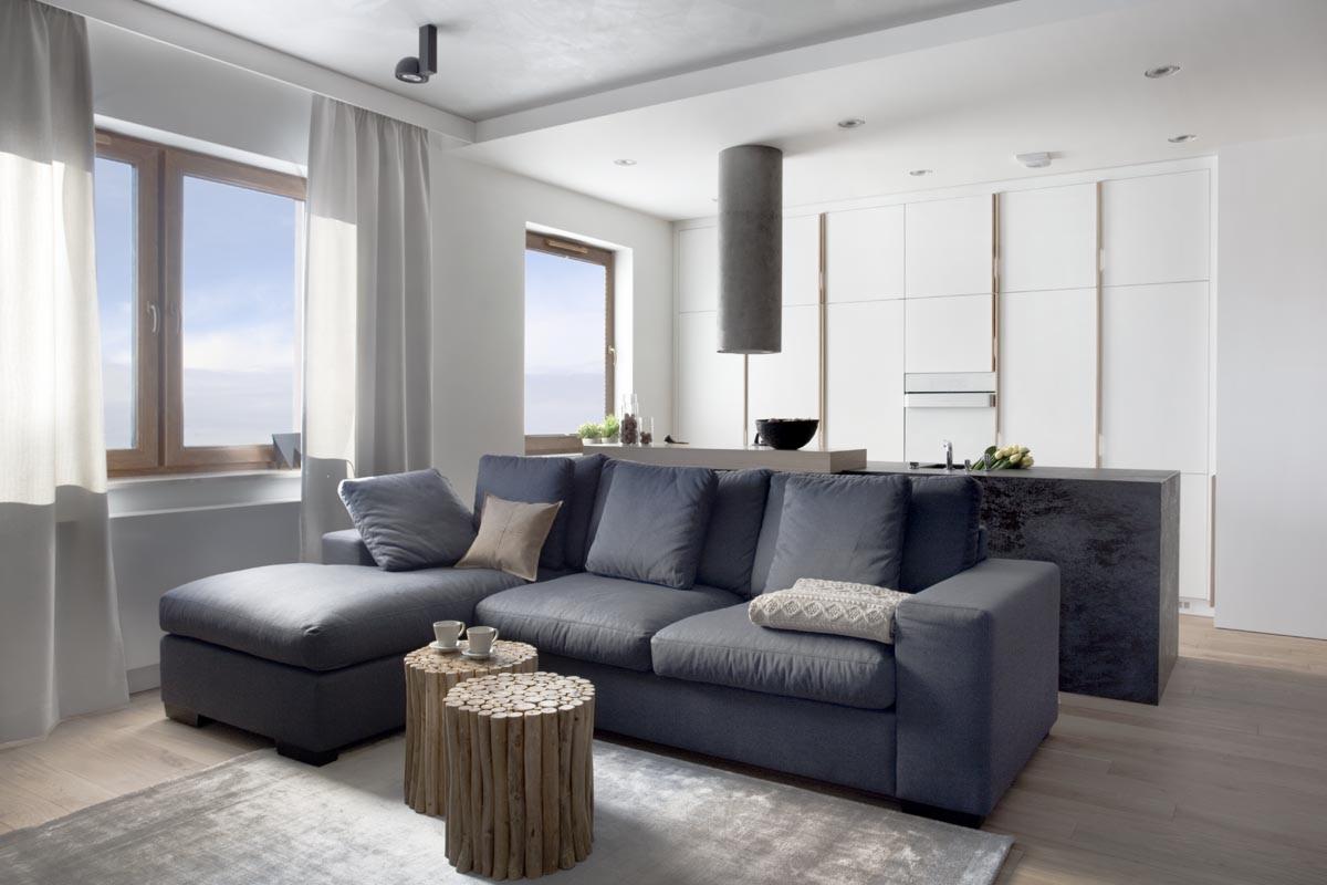 Nowoczesny salon z szarą sofą narożną, stolikami kawowymi z drewna, otwartą kuchnią z białymi szafkami i betonowym okapem, widok z okna na błękitne niebo.