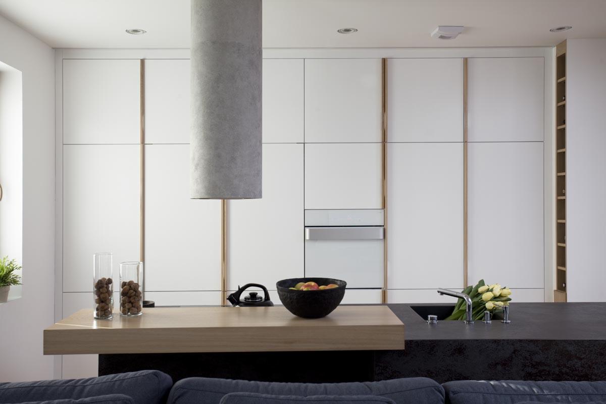 Minimalistyczna kuchnia z białymi szafkami, drewnianymi akcentami i cylindrycznym okapem. Na blacie drewnianym szklanki z brązowymi kulkami i czarna misa z owocami. W tle widać fragment szarej kanapy.