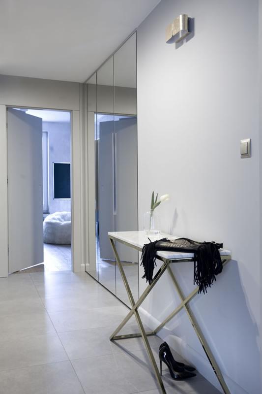 Nowoczesny przedpokój z lustrzaną szafą, marmurowym stolikiem konsolowym na złotym stelażu, białą lilią w wazonie i czarnymi szpilkami na podłodze.