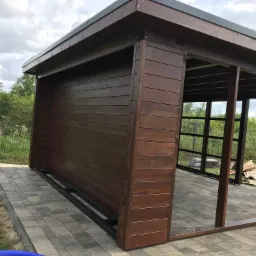 Drewniana konstrukcja zadaszenia w trakcie budowy, widoczne elementy szkieletowe i obudowa z ciemnego drewna, posadowiona na wybrukowanym tarasie, w tle zieleń ogrodu.