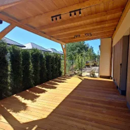 Zadaszony taras z desek, oświetlony punktowymi lampami, z widokiem na ogród i domy sąsiadów, otoczony tujami.