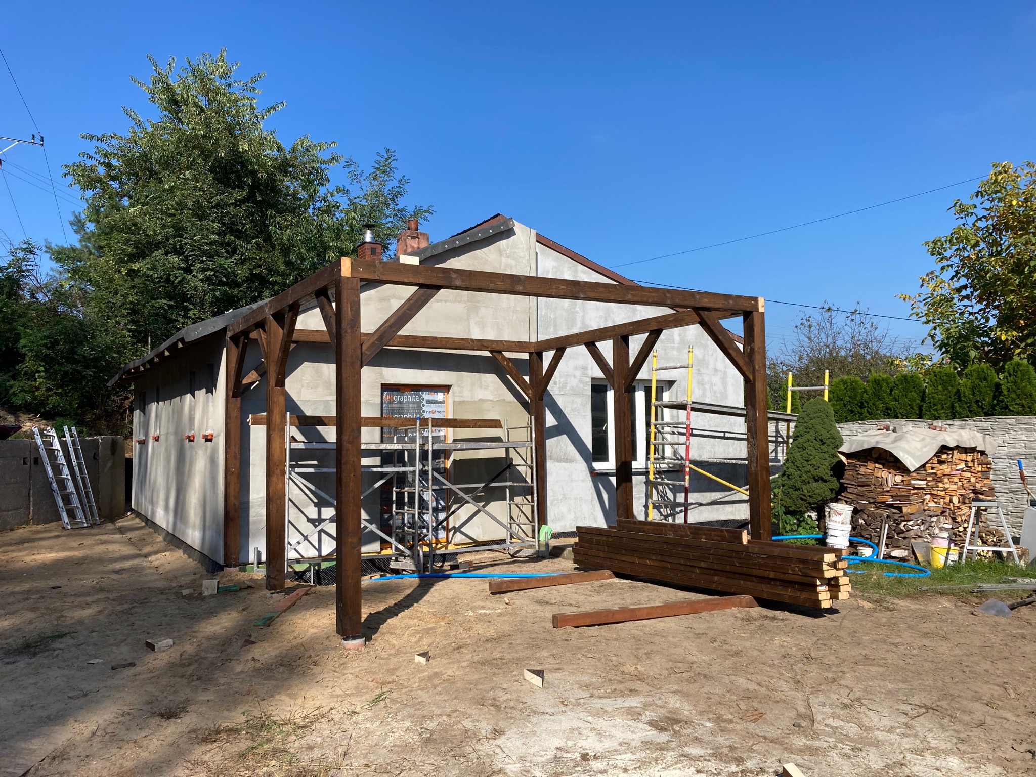 Wznoszona drewniana konstrukcja pergoli przy szarej elewacji domu, widoczne rusztowania i przygotowane drewno, błękitne niebo w tle.