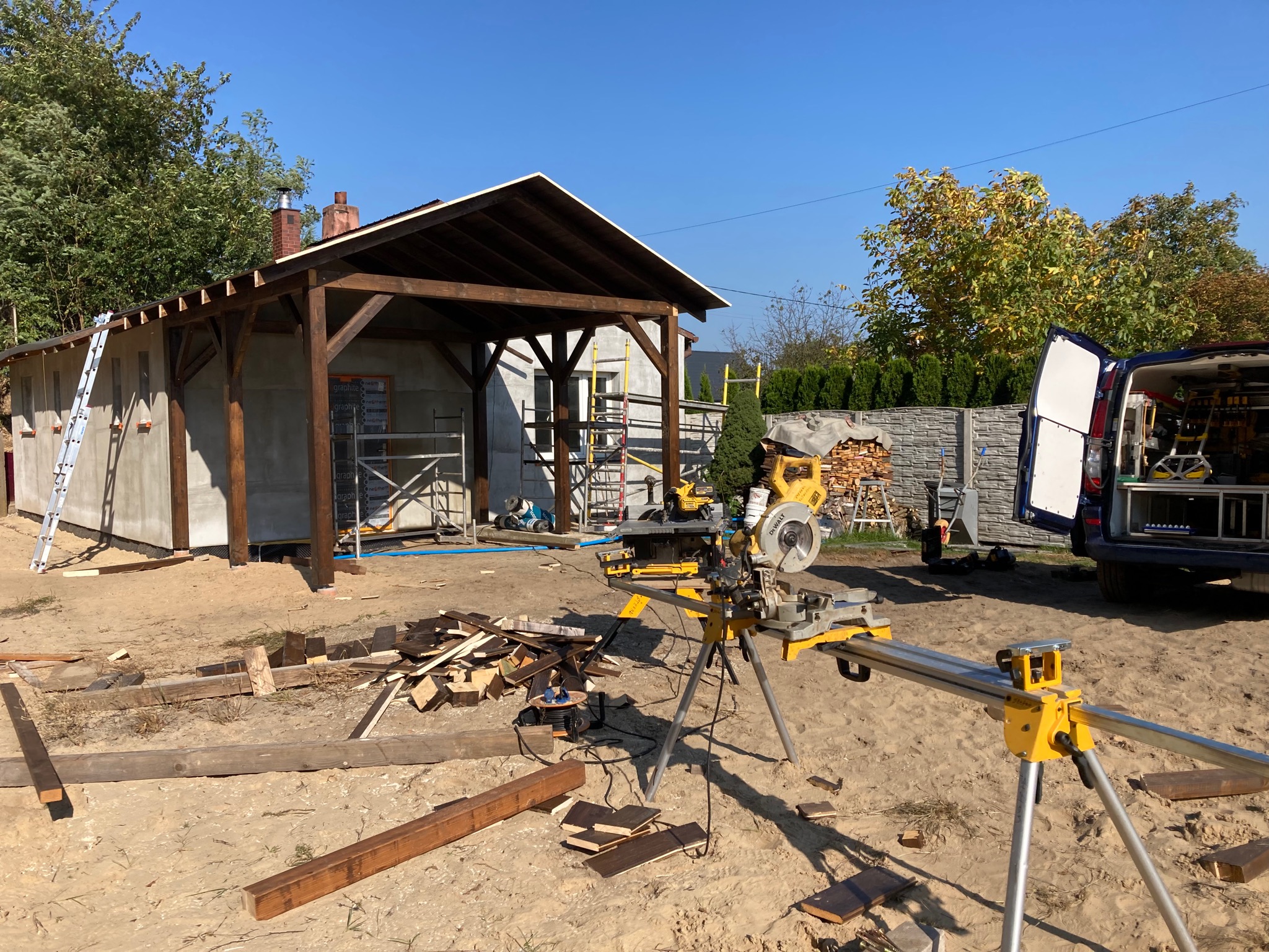 Wznoszenie drewnianej konstrukcji z zadaszeniem na piaszczystym podłożu, widoczne rusztowania, drabina oparta o ścianę budynku w tle, oraz piła stołowa na statywie i samochód dostawczy z otwartymi...