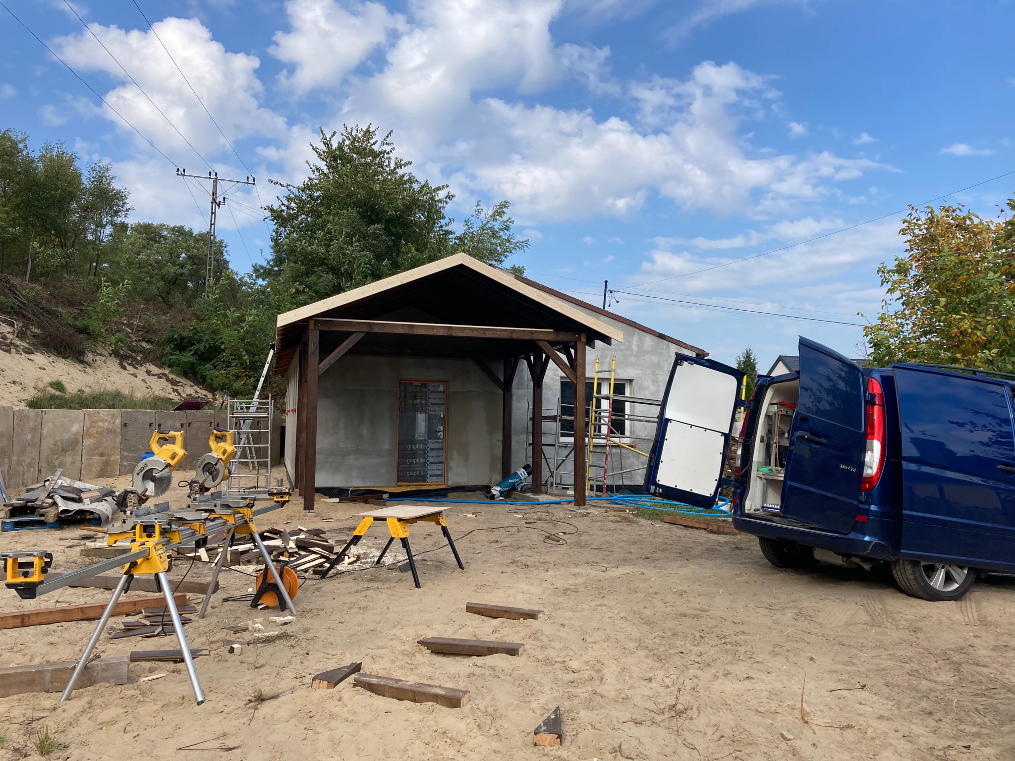 Budowa drewnianej konstrukcji z zadaszeniem na piaszczystym terenie, widoczne narzędzia stolarskie i otwarty samochód dostawczy w tle.