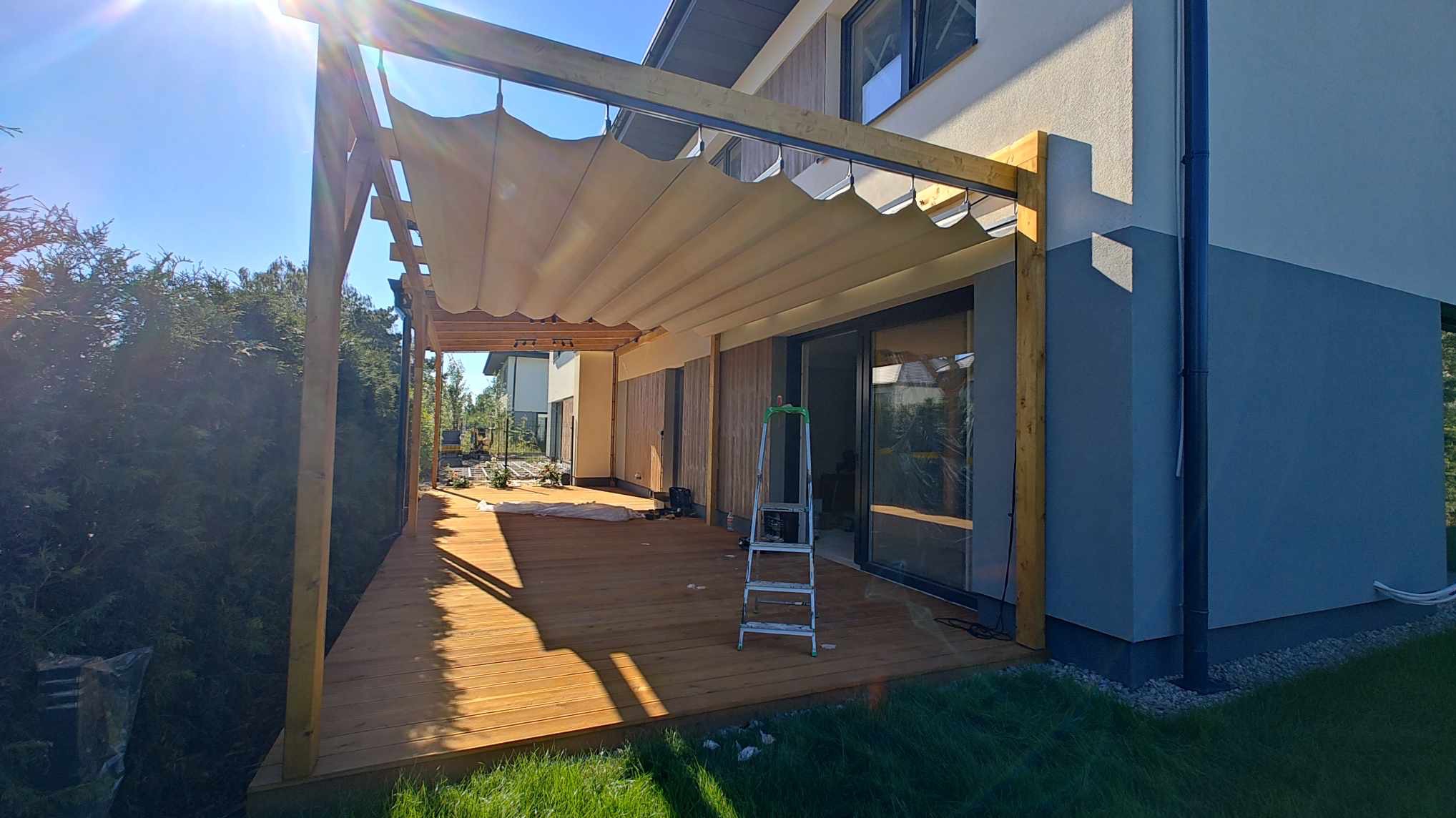Drewniany taras z pergolą z rozsuwanym zadaszeniem, częściowo zasłoniętym folią ochronną, drabina oparta o okno tarasowe, widok na ogród i dom w tle.